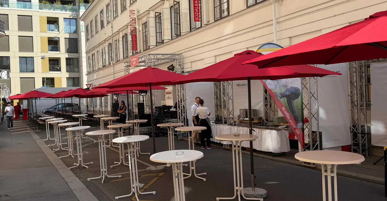 Weiße Stehtische und rote Sonnenschirme in einer Wiener Gasse bei einem Straßenfest
